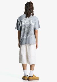 Grå kortærmet grafisk t-shirt med hvid tekst og detaljerede designs på ryggen. Sammen med hvide knælange shorts og gule sko.