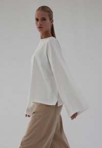 Luźna, biała bluzka z długim rękawem wykonana z bawełny, z okrągłym dekoltem typu crew, bocznymi rozcięciami i szerokimi rękawami, zestawiona z jasnobeżowymi spodniami.