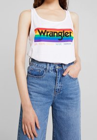 Vit ärmlös linne med en flerfärgad regnbågsdesign och "Wrangler"-text, i kombination med högmidjade blå denimjeans.