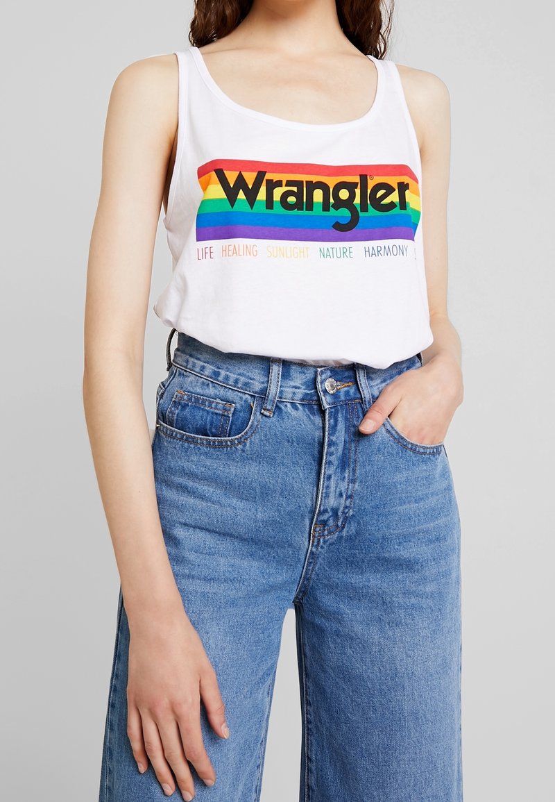 Vit ärmlös linne med en flerfärgad regnbågsdesign och "Wrangler"-text, i kombination med högmidjade blå denimjeans.