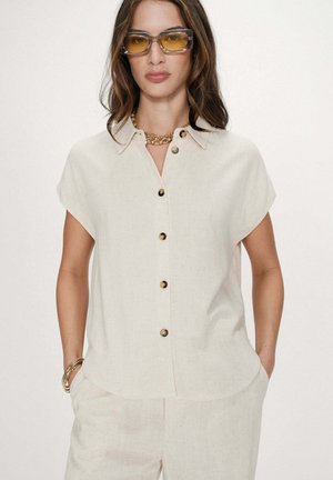 Femme portant une chemise à manches courtes boutonnée blanc cassé, pantalon assorti, lunettes de soleil teintées jaune, collier en chaîne dorée et bracelets, les mains dans les poches.
