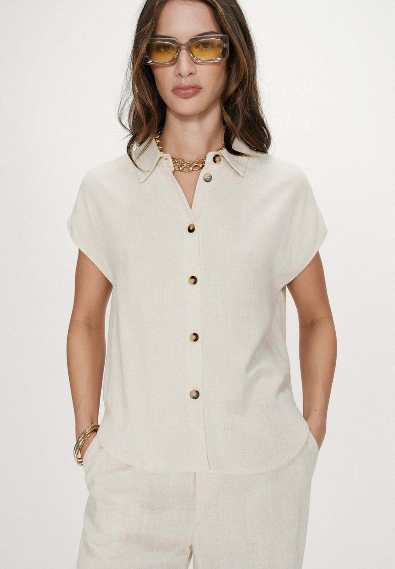 Mujer con camisa de manga corta color blanco roto abotonada, pantalones a juego, gafas de sol con lente amarilla, collar de cadena dorada y pulseras, con las manos en los bolsillos.