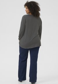 Blouse grise à manches longues avec une coupe décontractée et un pli dans le dos, portée avec un jean bleu foncé. Texture lisse, ourlet arrondi et détails subtils.