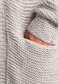 Maglione lavorato a maglia grigio chiaro con un motivo testurizzato e una grande tasca laterale, dotato di un polsino morbido e spesso a coste.