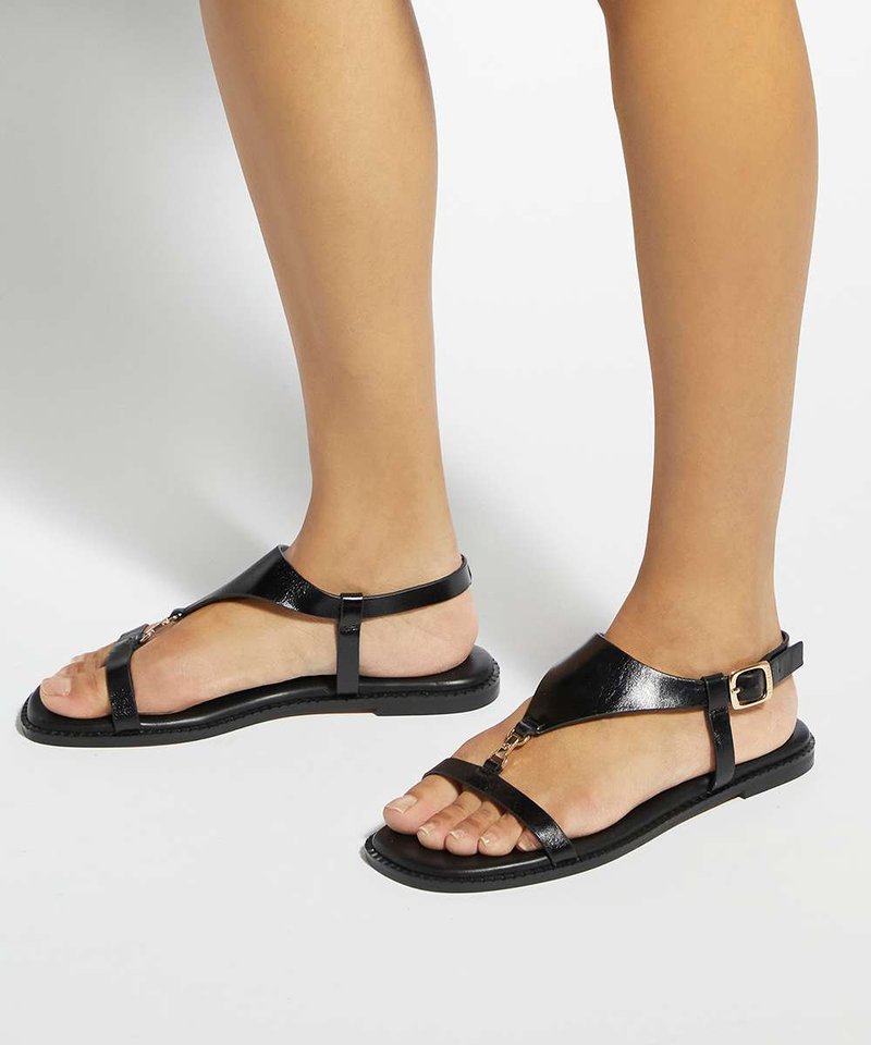 Pieds portant des sandales plates noires à fines lanières avec une boucle dorée sur un fond clair uni.