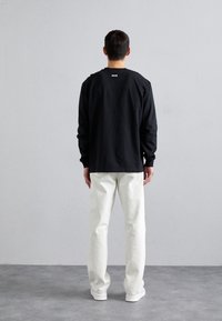 Sort langærmet skjorte med lille hvidt logo på ryggen, kombineret med hvide bukser med lige ben og hvide sneakers. Ensfarvet baggrund.