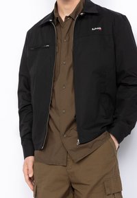 Black lightweight jacket with a zippered front, side pockets, and a chest pocket. Paired with a brown button-up shirt.