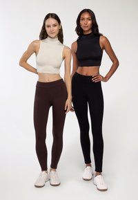 Dwie modelki noszą dopasowane bluzki bez rękawów oraz legginsy. Jedna bluzka jest jasnobeżowa, a druga czarna. Legginsy są brązowe i czarne, odpowiednio.