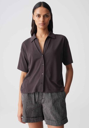 Donna con lunghi capelli scuri che indossa una camicia scura a maniche corte abbottonata e pantaloncini grigi a righe con orlo arrotolato su uno sfondo semplice.