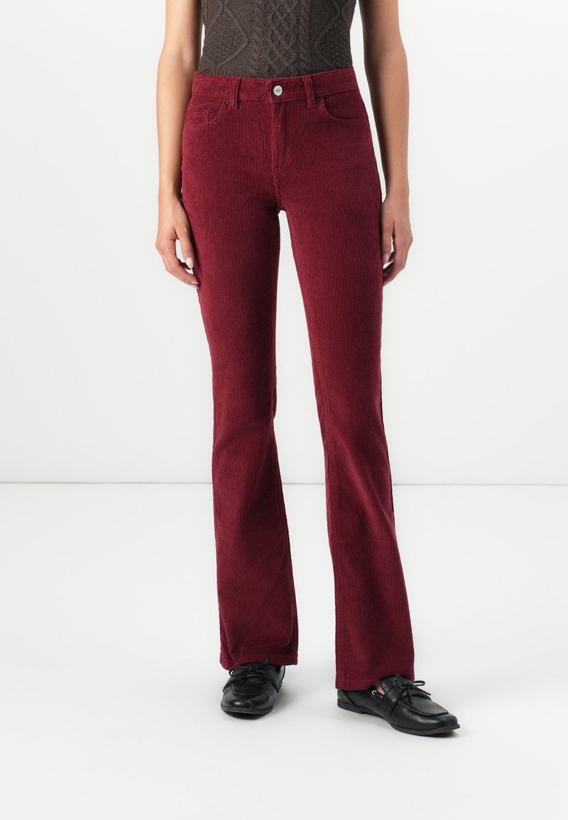 Personne portant un pantalon en velours côtelé bordeaux taille haute et des mocassins noirs, se tenant devant un fond blanc uni.