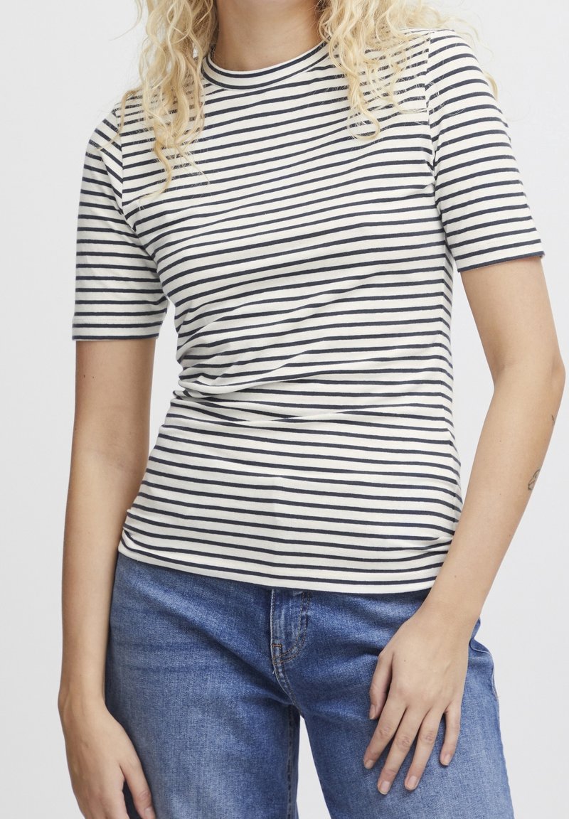 Femme portant un t-shirt à manches courtes blanc et noir à rayures, avec un jean bleu, debout avec une main posée sur la hanche.
