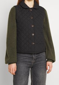 Gilet trapuntato nero con colletto tondo e bottoni marroni, abbinato a maniche lunghe in tessuto verde oliva. Indossato sopra jeans in denim scuro.