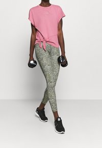 Rosa T-shirt med kort ärm och knut framtill, matchad med gröna leggings i djurtryck. Modellen håller hantlar och har på sig svarta träningkskor.