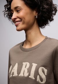 Brązowa bluza z napisem "PARIS" wykonanym w kremowym, teksturowanym materiale, z okrągłym dekoltem i prążkowanym kołnierzem. Modelka ma kręcone włosy i srebrne kolczyki.