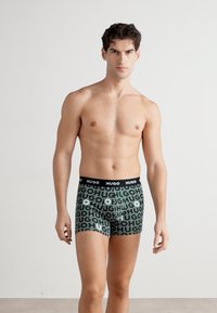Schwarze Taillengummis mit "HUGO"-Schriftzug; grüne Boxershorts mit wiederholtem "HUGO"-Grafikdesign in unterschiedlichen Größen; glatte Stofftextur.