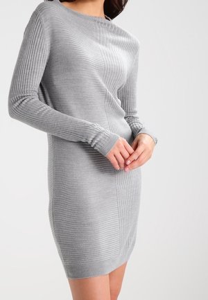 Woman in a fitted, long-sleeve light gray ribbed knit dress standing with hands gently clasped in front.