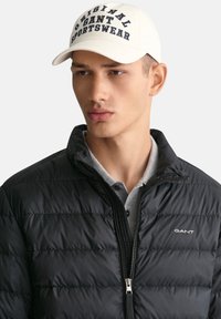 Sort pufferjakke med lynlås og GANT-logo; grå polo skjorte under; hvid baseballcap med marineblå tekst "Original GANT Sportswear."