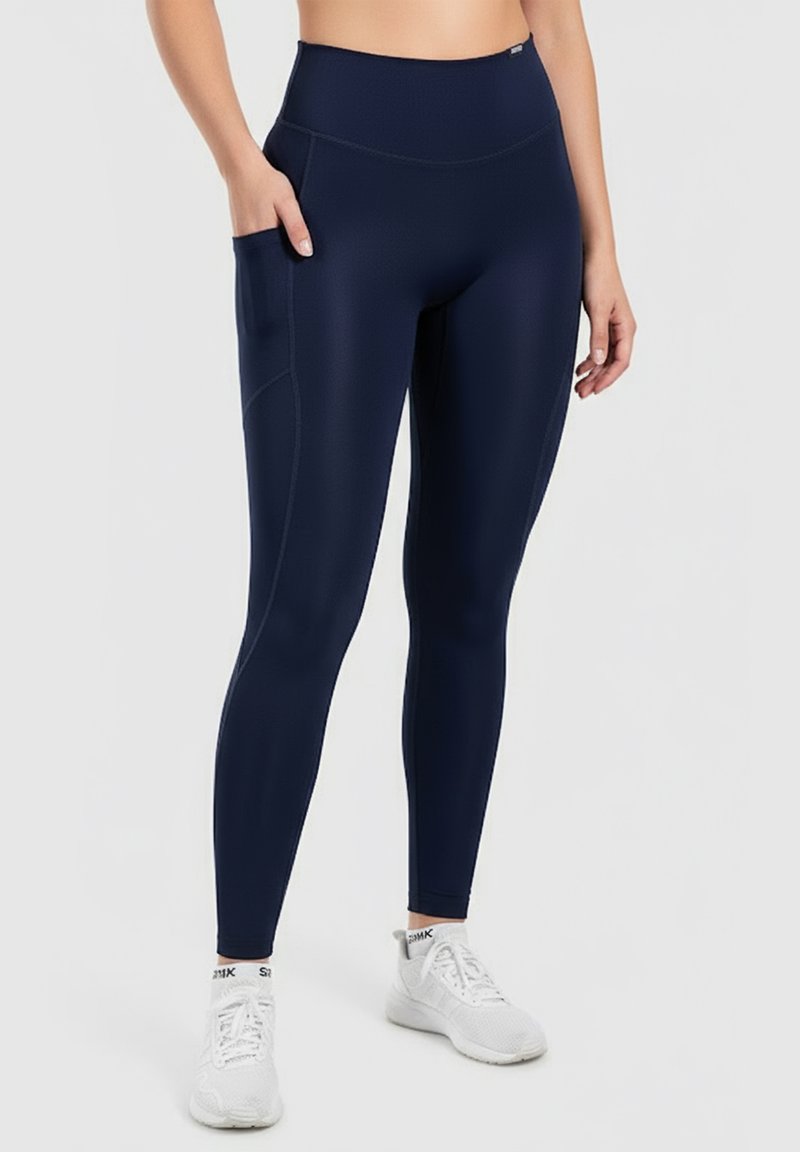 Marinblå högmidjade leggings med en sidficka, slät textur och åtsittande design. Matchat med vita träningsskor.