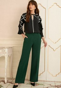 Blusa nera con dettagli a ruffle bianchi e apertura a chiave, abbinata a pantaloni larghi verdi e décolleté neri. Ambiente caratterizzato da texture.