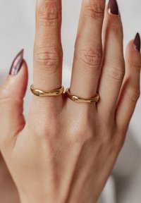 Anelli color oro con bordi lisci e arrotondati indossati su due dita. La pelle della mano, con una consistenza naturale, è visibile.