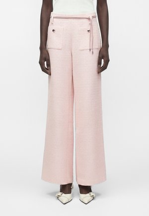 Femme portant un pantalon rose taille haute à jambes larges avec poches plaquées à l'avant et détails de boutons, associé à des talons blancs à bout pointu.