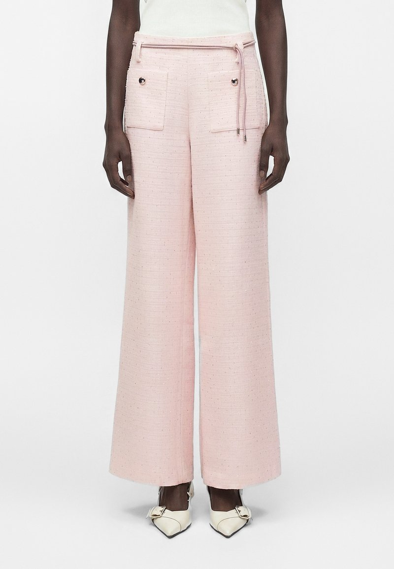 Γυναίκα φοράει ροζ παντελόνια με ψηλή μέση και φαρδιά ποδάρια, με εμπρόσθιες τσέπες και λεπτομέρειες με κουμπιά, συνδυασμένα με λευκές μυτερές γόβες.