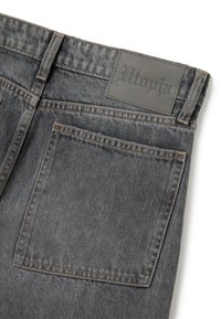 Jeans in denim grigio con una patch in pelle etichettata "Utopia." Presentano cuciture a contrasto e una tasca posteriore. Tessuto texture con una finitura scolorita.