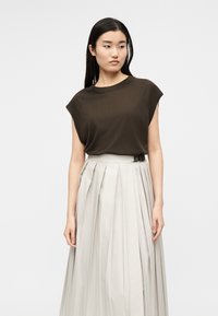 Γυναίκα με μακριά μαύρα μαλλιά που φοράει σκούρο καφέ αμάνικη μπλούζα και ανοιχτό μπεζ πλισέ φούστα με ψηλή μέση και μαύρη ζώνη με αγκράφα.