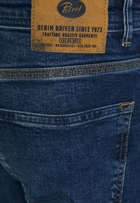 Jeans i denim med en mörkblå ton, utrustade med en beige läderetikett, markerad sömnad och en klassisk design med fem fickor.