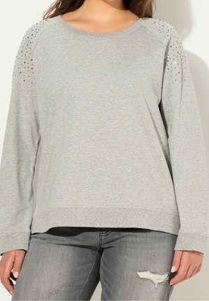 Kobieta ubrana w jasnoszary top z długim rękawem z małymi ozdobami na ramionach oraz podarte szare dżinsy, stojąca na jednolitym tle.