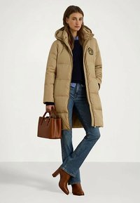 Sieviete, tērpusies bēšā garā pildījuma mētelī, zilu džinsu biksēs, brūnās papēžu zābakos, turot brūnu ādas rokassomu, stāv uz neitrālas fona.