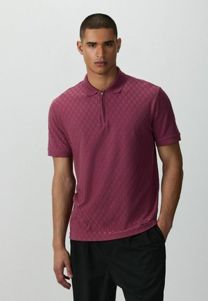 Jonge man met kort haar die een gemêleerd bordeauxrood poloshirt met korte mouwen en een zwarte broek draagt, met één hand in zijn zak.