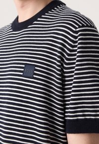 Gros plan d'une personne portant un t-shirt à manches courtes rayé bleu marine et blanc avec un col bleu marine et un écusson au logo BOSS sur la poitrine.