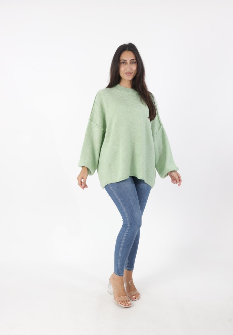 Hellgrüner, übergroßer Strickpullover mit hohem Kragen, überschnittenen Schultern und abgerundetem Saum, kombiniert mit schmal geschnittenen blauen Jeans und transparenten Absätzen.