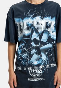 Čierne tričko s modrou bleskovou grafikou, veľkou hviezdou obsahujúcou lebku a výrazným logom a textom „DCSHOECOUSA“ na prednej strane.