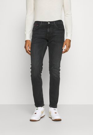 Osoba ubrana w czarne obcisłe dżinsy i białe trampki z beżowymi akcentami, stojąca na jednolitym jasnym tle.