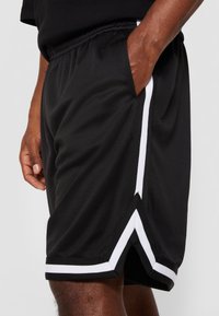 Homme portant un short de sport noir avec des bandes blanches, main dans la poche, montré de la taille jusqu'au milieu de la cuisse sur un fond uni.