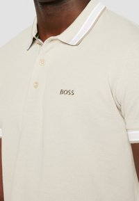 Gros plan d'une personne portant un polo beige avec des bordures blanches sur le col et les manches, avec le mot "BOSS" brodé sur la poitrine.