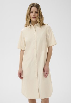Kobieta w beżowej, sięgającej kolan sukience koszulowej z krótkim rękawem, stojąca na jednolitym tle.