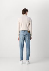Haut à manches longues beige clair associé à un jean bleu délavé. Le jean a une coupe droite avec une étiquette en cuir marron sur la ceinture arrière. Baskets blanches.