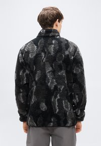 Μαύρο και γκρι fleece μπουφάν με μαλακή, υφή επιφάνεια. Διαθέτει ψηλό γιακά και άνετη εφαρμογή. Η πλάτη εμφανίζει ένα κηλιδωτό μοτίβο.