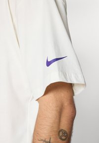 White cotton t-shirt with short sleeves featuring a purple Nike logo on the left sleeve. Simple design and smooth texture.