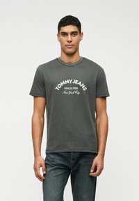 Γκρίζο βαμβακερό μπλουζάκι με στρογγυλή λαιμόκοψη, κοντά μανίκια και λογότυπο "TOMMY JEANS" με το κείμενο "SINCE 1985" και "New York City" σε άσπρο χρώμα.