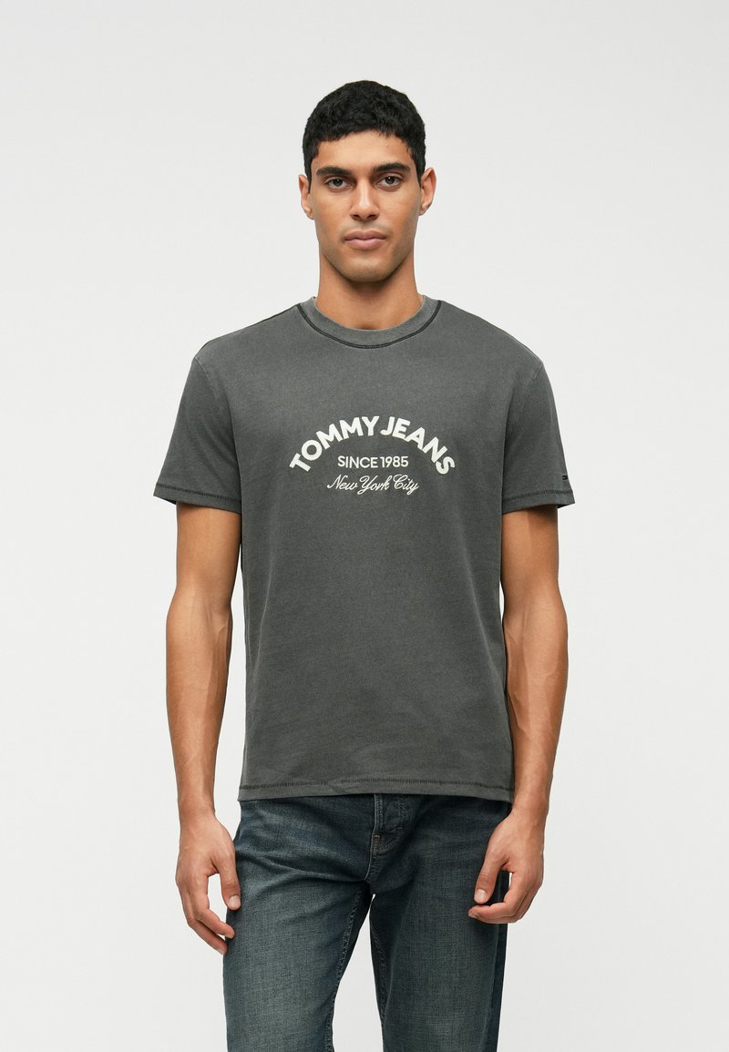 Γκρίζο βαμβακερό μπλουζάκι με στρογγυλή λαιμόκοψη, κοντά μανίκια και λογότυπο "TOMMY JEANS" με το κείμενο "SINCE 1985" και "New York City" σε άσπρο χρώμα.
