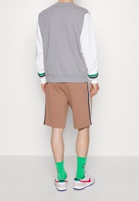 Manlig figur sedd bakifrån, iförd en grå och vit sweatshirt, bruna shorts, ljusgröna strumpor och sneakers, mot en enkel bakgrund.