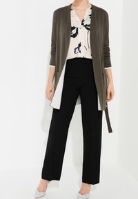 Cardigan vert olive sur une blouse à fleurs, associé à un pantalon noir taille haute et des talons gris clair. Le cardigan présente un design à devant ouvert.