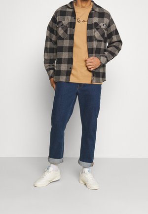 Mand iført beige t-shirt, sort- og gråternet skjorte med knapper, blå opkørte jeans, hvide sokker og hvide sneakers, står mod en hvid baggrund.