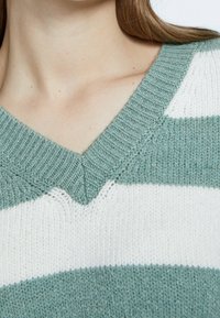 Sweter dzianinowy z dekoltem w kształcie litery V, z poziomymi paskami w delikatnym zielonym i białym odcieniu, z wyraźnie zaznaczoną strukturą ściągacza i detalami przy dekolcie.