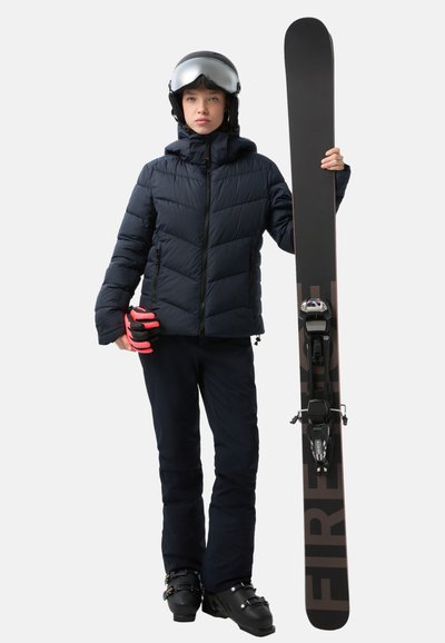 Gepolsterte marineblaue Jacke, schwarze Skihosen und Skischuhe. Hält ein schwarzes Snowboard mit "FIRE"-Druck und pinken Handschuhen. Silberner Helm mit Visier.