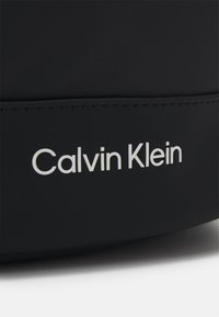 Černý textilní doplněk s hladkým povrchem, s viditelným bílým logem "Calvin Klein" výrazně vytištěným. Jednoduchý design bez ozdob.